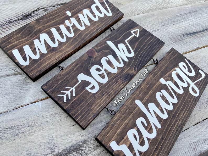 May include: Three wooden signs with white lettering hanging from a string. The top sign reads "unwind", the middle sign reads "soak" with an arrow pointing to the right, and the bottom sign reads "recharge".