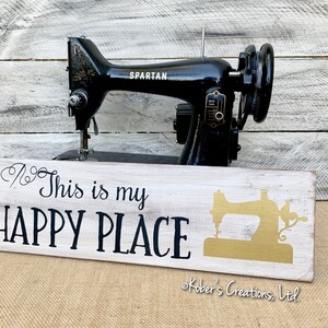 May include: White wooden sign with black text that reads "This is my HAPPY PLACE" and a gold silhouette of a sewing machine. The sign is sitting in front of a black vintage sewing machine.