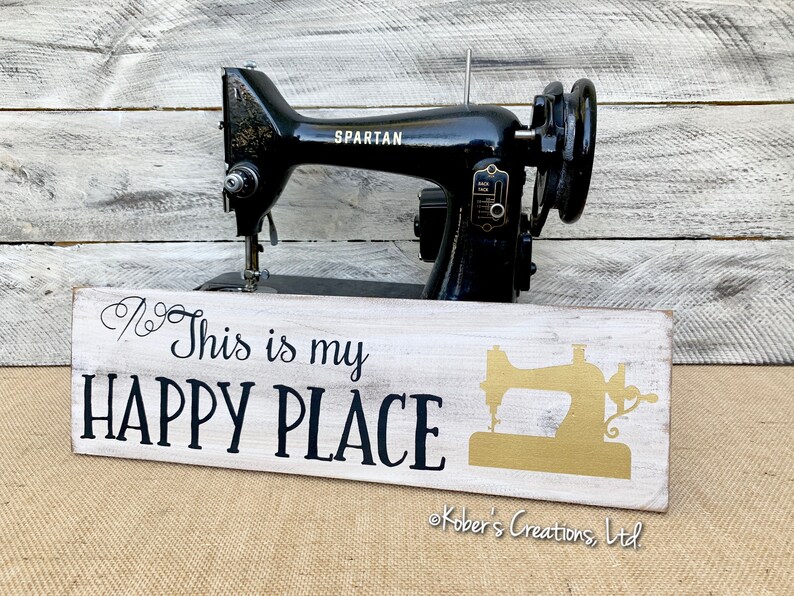 May include: White wooden sign with black text that reads "This is my HAPPY PLACE" and a gold silhouette of a sewing machine. The sign is sitting in front of a black vintage sewing machine.