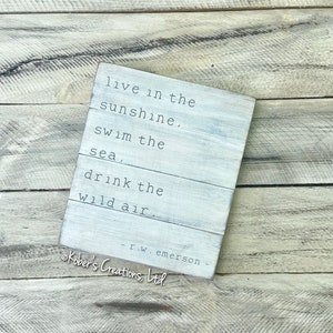 May include: White wooden sign with a quote by R.W. Emerson: "live in the sunshine, swim the sea, drink the wild air". The sign is on a woodgrain background.