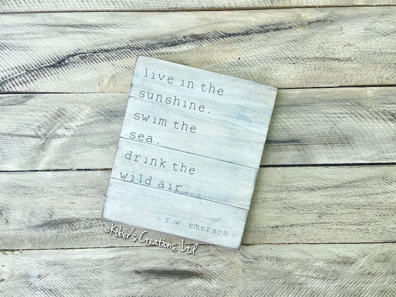 May include: White wooden sign with a quote by R.W. Emerson: "live in the sunshine, swim the sea, drink the wild air". The sign is on a woodgrain background.