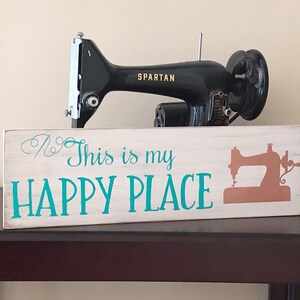 May include: White wooden sign with the text "This is my HAPPY PLACE" in teal blue lettering. A silhouette of a sewing machine is on the right side of the sign. A black vintage sewing machine is sitting on top of the sign.
