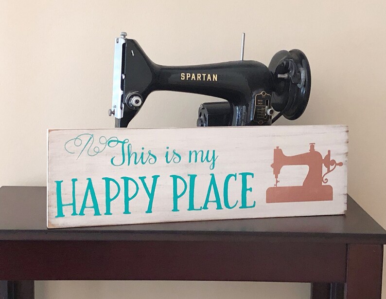 May include: White wooden sign with the text "This is my HAPPY PLACE" in teal blue lettering. A silhouette of a sewing machine is on the right side of the sign. A black vintage sewing machine is sitting on top of the sign.