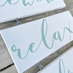 May include: White wooden sign with the word "relax" written in a light blue cursive font. The sign is hanging from two silver hooks.