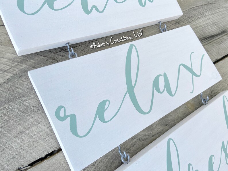 May include: White wooden sign with the word "relax" written in a light blue cursive font. The sign is hanging from two silver hooks.