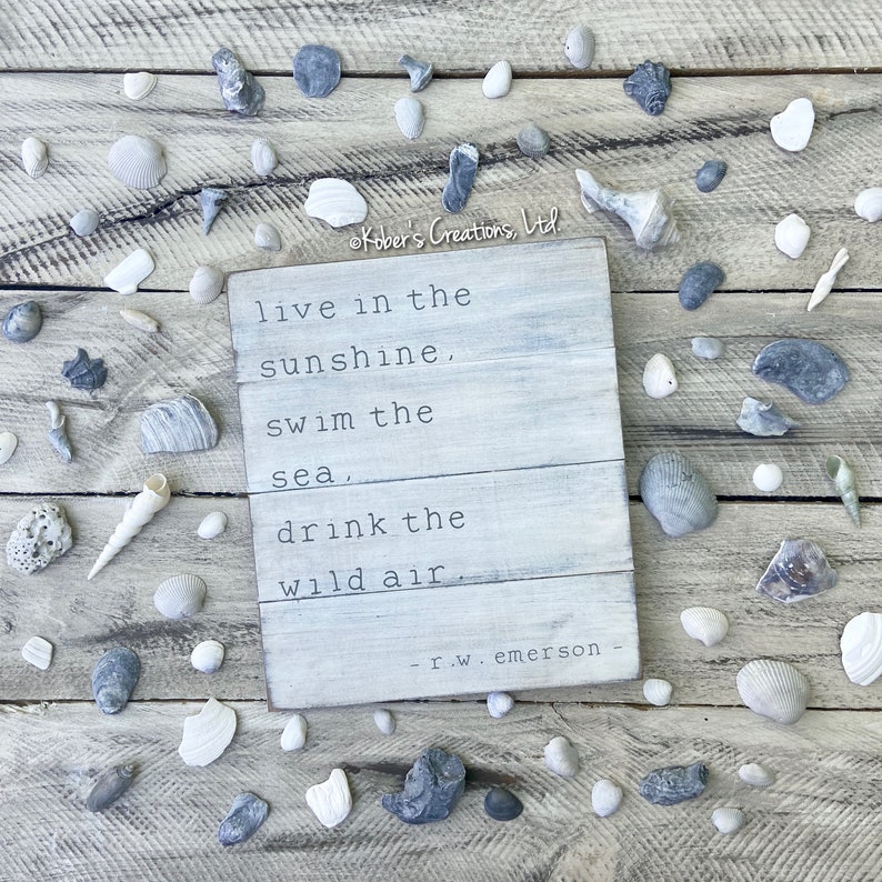 May include: White wooden sign with a quote by R.W. Emerson: "live in the sunshine, swim the sea, drink the wild air". The sign is surrounded by seashells and stones on a wooden background.