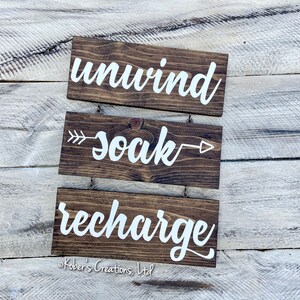 May include: Three wooden signs with white lettering hanging on a light wood background. The top sign says "unwind", the middle sign says "soak" with an arrow pointing to the right, and the bottom sign says "recharge".