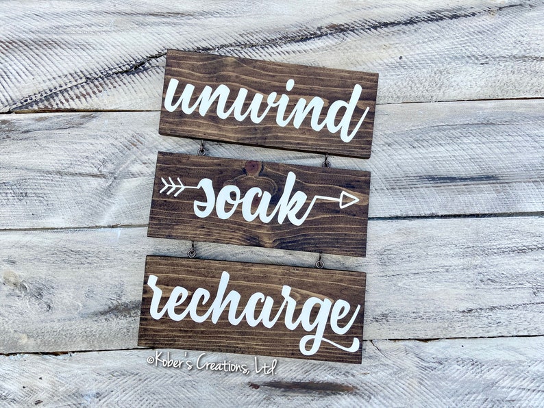 May include: Three wooden signs with white lettering hanging on a light wood background. The top sign says "unwind", the middle sign says "soak" with an arrow pointing to the right, and the bottom sign says "recharge".