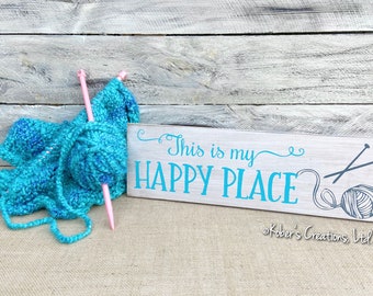 May include: A wooden sign with the text "This is my happy place" in teal blue lettering. The sign also features a gray yarn ball and knitting needles. A teal blue yarn ball and pink knitting needles are in the foreground.