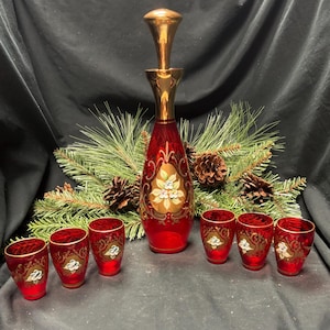 May include: A red glass decanter set with gold detailing and floral patterns. The set includes a tall decanter with a gold stopper and six matching shot glasses. The set is decorated with gold floral patterns and is suitable for serving spirits.