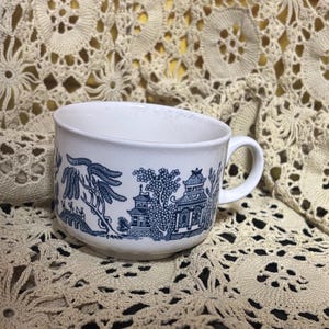 Puede incluir: Taza de té de cerámica blanca con un diseño azul de patrón Willow. La taza presenta un asa y una escena de paisaje detallada con árboles, puentes y edificios. La taza está sobre un fondo de tela crema de tejido abierto.