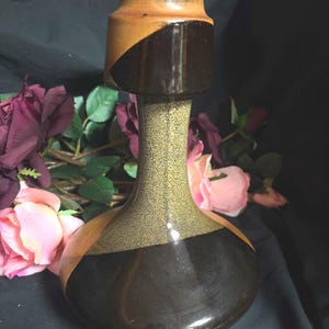 Puede incluir: Un jarrón de cerámica con base bulbosa y cuello estrecho. La superficie presenta un patrón de esmalte tricolor con un acabado marrón oscuro brillante en la parte inferior y superior, separado por una banda central de esmalte verde oliva texturizado.