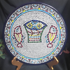 May include: A round decorative plate featuring a mosaic design with a central basket motif flanked by two fish. The background is white with black grid lines, surrounded by a dark blue border with intricate white, red, yellow, and blue patterns.