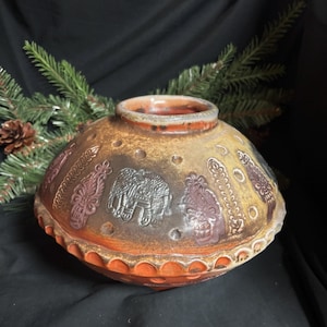May include: A handcrafted ceramic vase with a wide, rounded body and a small opening. The vase features embossed elephant and floral designs in shades of brown, red, and gray. The base and rim have a textured, orange-brown finish.