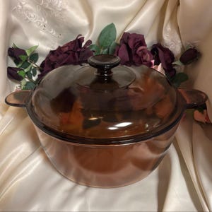 May include: A round, amber-coloured glass casserole dish with a matching transparent lid. The pot features two integrated side handles and a central knob on the lid. The glass is translucent, allowing light to pass through the warm brown material.