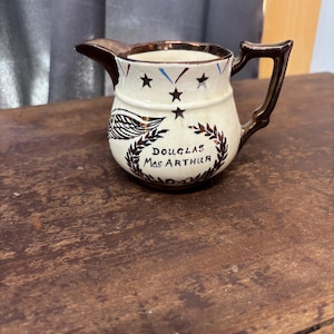 May include: A small, vintage cream pitcher with a brown handle and rim. The pitcher features a design with stars, wings, and the text "DOUGLAS MAC ARTHUR" in a decorative font. The pitcher is on a wooden surface.