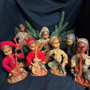 May include: A collection of seven figurines depicting characters in pointed hats and tunics, each holding a small stringed instrument. The figures are dressed in red, tan, and grey clothing, with painted facial features and seated on textured bases.