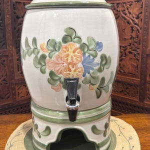 May include: A white ceramic beverage dispenser with a floral design in shades of orange, pink, and blue. The dispenser has a green rim and base, a black spigot, and a matching lid. The dispenser is on a round, beige placemat.