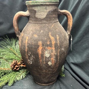 Puede incluir: Un jarrón rústico de cerámica con dos asas, con exterior marrón oscuro y vetas anaranjadas. El borde es verde y el jarrón tiene un aspecto envejecido. Una rama de pino y una piña están en primer plano.