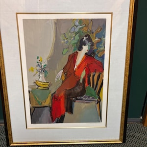 May include: Framed art print featuring a seated figure in a red dress. The artwork has a gold and black frame, with a white mat. The print depicts a woman seated in a chair, with a table and flowers to the left. The style is impressionistic.