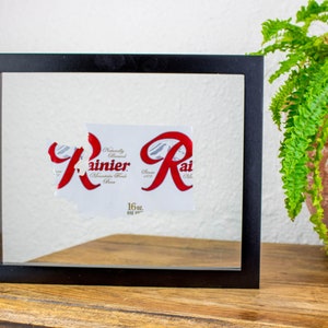 Puede incluir: Una obra enmarcada con una etiqueta rasgada que muestra la palabra "Rainier" en escritura roja. La etiqueta está sobre un fondo blanco dentro de un marco negro. Un helecho en maceta está a la derecha sobre una superficie de madera.