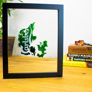 Könnte beinhalten: Ein gerahmtes Kunstwerk mit einer grünen und weißen Karte von Dänemark, mit dem Carlsberg-Logo. Das Kunstwerk ist in einem schwarzen Rahmen gefasst. Bücher und eine Pflanze sind im Hintergrund zu sehen.