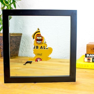 May include: A framed artwork featuring a yellow map of England with a beer-themed design. The artwork includes the text "JB AL" and "1 PINT". The frame is black, and the artwork is displayed on a wooden surface.