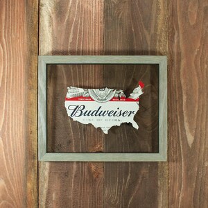 May include: A framed artwork featuring a cutout of the United States map, crafted from a Budweiser beer label. The label's red, white, and silver design is visible, with the brand name prominently displayed. The frame is a muted gray, set against a wooden background.