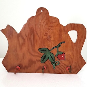 Op de afbeelding: Houten sleutelhouder in de vorm van een theepot. Het bruine hout heeft een geschilderd aardbeienontwerp met groene bladeren en een rode bes. Onder het aardbeienontwerp bevinden zich drie metalen haken.