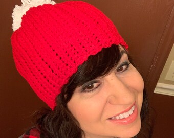 May include: A handmade red crochet hat with a white pom-pom. The hat is a beanie style, with a ribbed texture. The person in the image is wearing a red and black plaid shirt and a gray shirt.