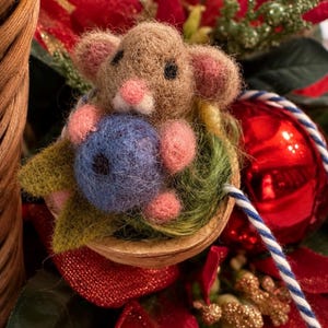 May include: A needle-felted tan mouse with pink ears and nose holding a blue berry, nestled inside a walnut shell with green felt leaves. The mouse has small black eyes and is positioned among red poinsettia petals and a blue and white string.