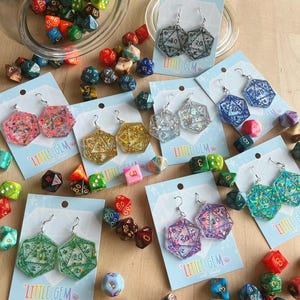 May include: A collection of colorful dice and several pairs of geometric resin earrings. The earrings are in various colors, including pink, gold, blue, and green, with the number 20 visible. The earrings are on light blue cards with the text "LITTLE GEM Craft Studio".