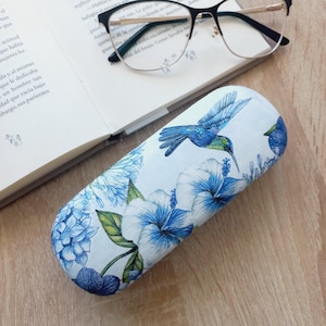 Könnte beinhalten: Ein weißes Brillenetui mit einem blauen Kolibri- und Blumenmuster. Das Etui zeigt einen lebendigen blauen Kolibri mit grünen Akzenten, umgeben von blauen Hibiskusblüten und grünen Blättern. Eine Brille liegt auf einem offenen Buch.