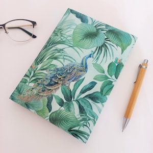 Può includere: Un taccuino rettangolare con copertina verde chiaro caratterizzato da un motivo dettagliato di pavone e foglie tropicali, accompagnato da un paio di occhiali con montatura sottile scura e una penna in legno marrone chiaro con clip e punta in metallo argentato.