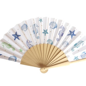 Può includere: Ventaglio bianco con manico in legno, con un design nautico. Il tessuto del ventaglio è decorato con stelle marine blu, cavallucci marini e conchiglie, con dettagli dorati e strisce beige. Il ventaglio è aperto, mostrando la sua forma curva.