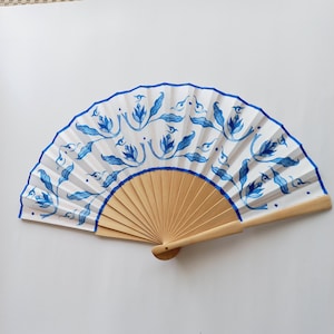 Op de afbeelding: Een handventilator met een witte achtergrond en een blauw bloemenpatroon. Het frame van de ventilator is gemaakt van natuurlijk hout en de randen zijn blauw omlijnd. De ventilator is open en toont het gedetailleerde patroon.