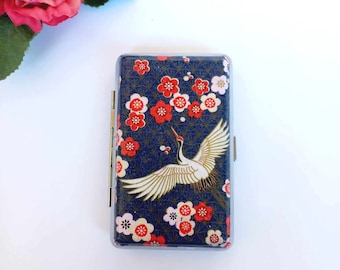 May include: A blue cigarette case with a white crane flying over a pattern of red and white flowers. The case has a silver hinge.