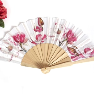 May include: A white folding fan with a wooden handle and a floral design featuring pink and white magnolia flowers.