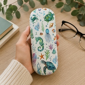 May include: A hard-shell eyeglasses case with a white background featuring a colorful watercolor print of various marine life including sea turtles, a seahorse, a crab, jellyfish, coral, seaweed, and seashells scattered across the surface.
