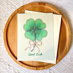 Pu&ograve; includere: Un biglietto di auguri con un'illustrazione di un quadrifoglio, un piccolo coniglietto bianco e la scritta "Good Luck". Il biglietto &egrave; su un vassoio di legno con una busta bianca.