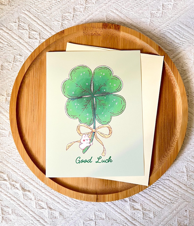 Pu&ograve; includere: Un biglietto di auguri con un'illustrazione di un quadrifoglio, un piccolo coniglietto bianco e la scritta "Good Luck". Il biglietto &egrave; su un vassoio di legno con una busta bianca.