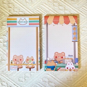 May include: Two illustrated notepads with cute animal characters. One notepad features a bear and a cat at a shop called "Meow Mart" with the word "OPEN". The other notepad shows a bear, cat, and rabbit at a shop with the text "ONE ORE ORE THREE 500 SIX".