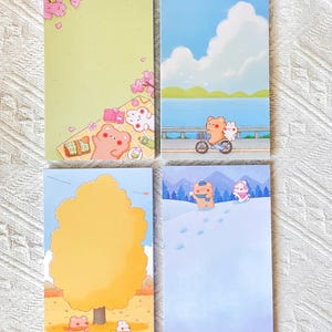 May include: Four rectangular memo pads featuring seasonal illustrations of a bear and a rabbit. The designs depict a spring picnic, a summer bike ride, an autumn tree scene, and a winter snow landscape with the characters playing in the snow.