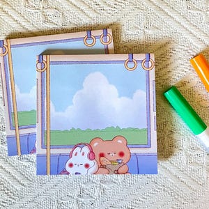 May include: Two square notepads with a cartoon illustration of a bear and bunny on a train, with a blue sky background. The notepads have gold-colored rings at the top. Two markers are visible in the background.