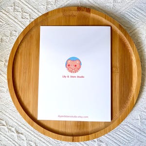 Pu&ograve; includere: Un biglietto di auguri bianco con un'illustrazione di orso dei cartoni animati, posto su un vassoio rotondo di legno. Il biglietto presenta il testo "Lily & Stars Studio" e l'indirizzo del sito web. L'orso &egrave; rosa con un cerchio blu.