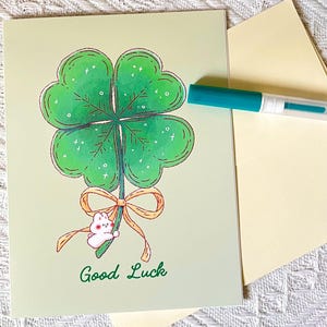 Pu&ograve; includere: Biglietto di auguri con un'illustrazione di un quadrifoglio verde con contorno marrone. Un piccolo coniglietto bianco tiene un fiocco. Le parole "Good Luck" sono scritte in verde. Sono presenti anche un pennarello turchese e una busta gialla.