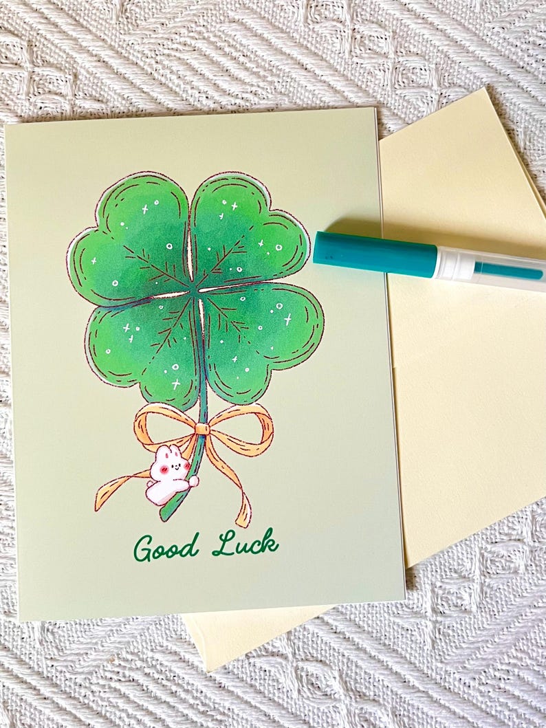 Pu&ograve; includere: Biglietto di auguri con un'illustrazione di un quadrifoglio verde con contorno marrone. Un piccolo coniglietto bianco tiene un fiocco. Le parole "Good Luck" sono scritte in verde. Sono presenti anche un pennarello turchese e una busta gialla.
