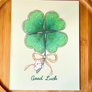 Pu&ograve; includere: Biglietto di auguri con un quadrifoglio verde. Un piccolo coniglietto bianco tiene lo stelo del quadrifoglio, legato con un nastro giallo. Le parole "Good Luck" sono scritte sotto.