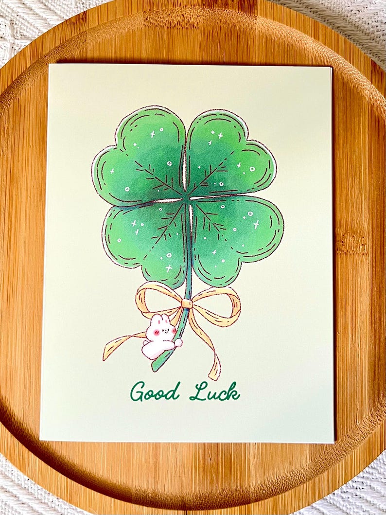 Pu&ograve; includere: Biglietto di auguri con un quadrifoglio verde. Un piccolo coniglietto bianco tiene lo stelo del quadrifoglio, legato con un nastro giallo. Le parole "Good Luck" sono scritte sotto.