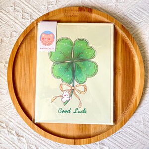 Pu&ograve; includere: Un biglietto di auguri con un quadrifoglio verde, un coniglietto bianco e un fiocco. Le parole "Good Luck" sono scritte in verde. Il biglietto &egrave; su uno sfondo verde chiaro ed &egrave; in una custodia di plastica trasparente. &Egrave; attaccata una piccola etichetta con un orsetto dei cartoni animati.